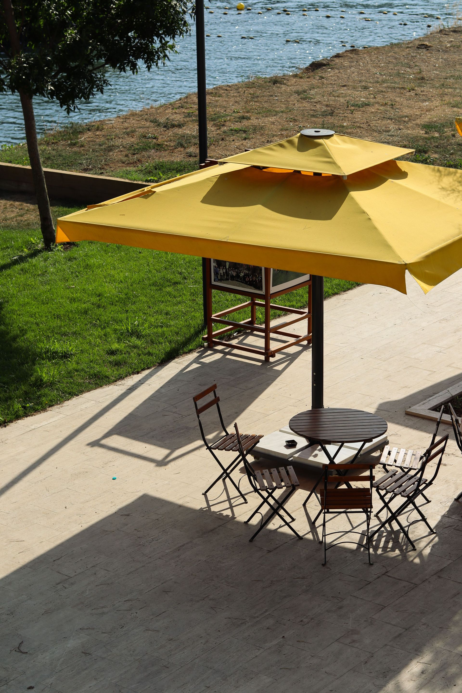 Serene outdoor cafe seating with a vibrant yellow umbrella by a riverside path.